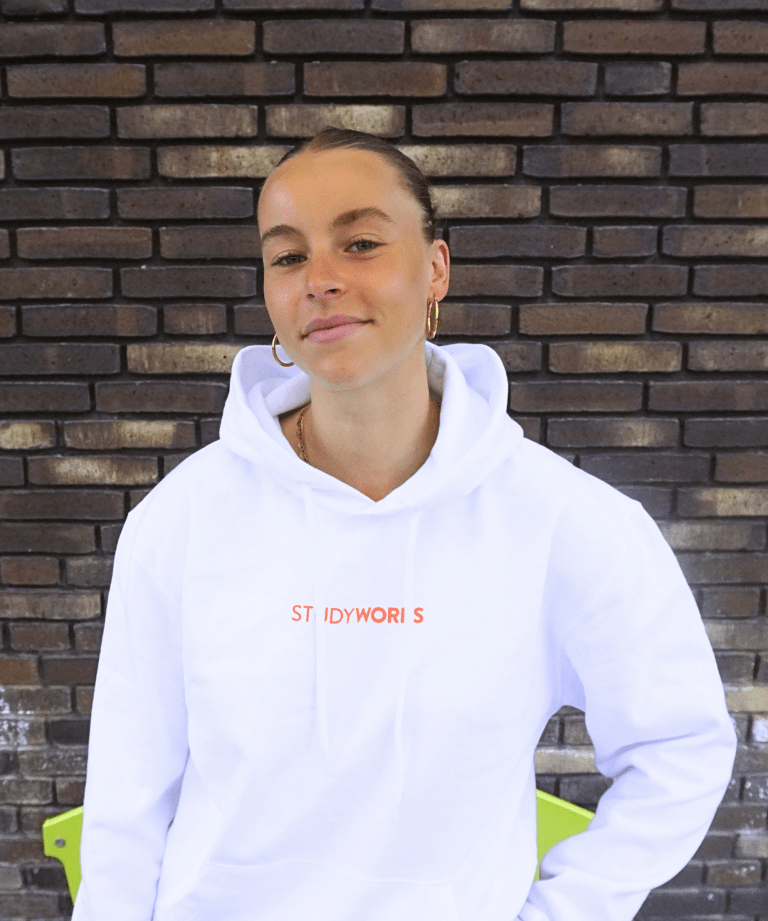 Fleurtje, pedagogisch medewerker bso via StudyWorks, in een witte StudyWorks-hoodie voor een bakstenen muur