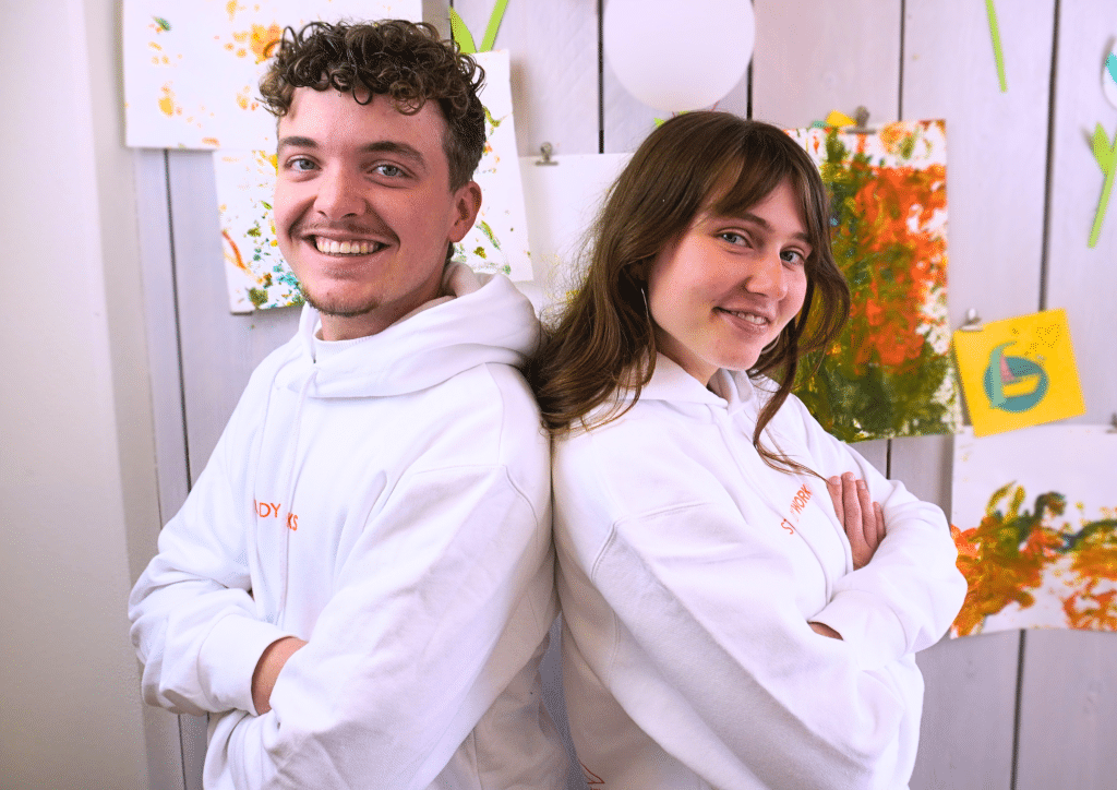 Twee lachende medewerkers van StudyWorks in witte hoodies staan rug aan rug, met kleurrijke kindertekeningen op de achtergrond.
