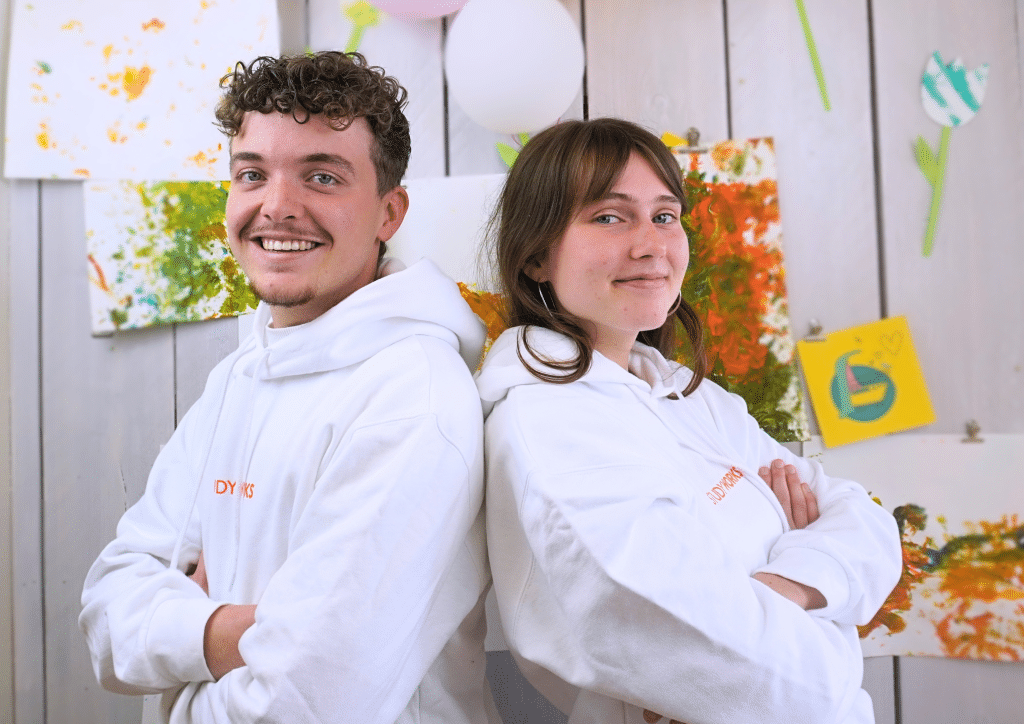 Twee lachende StudyWorkers in witte StudyWorks-hoodies staan rug aan rug voor een houten wand met kindertekeningen