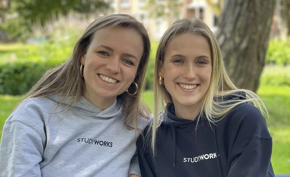 Twee StudyWorkers in StudyWorks-hoodies zitten lachend naast elkaar in het park